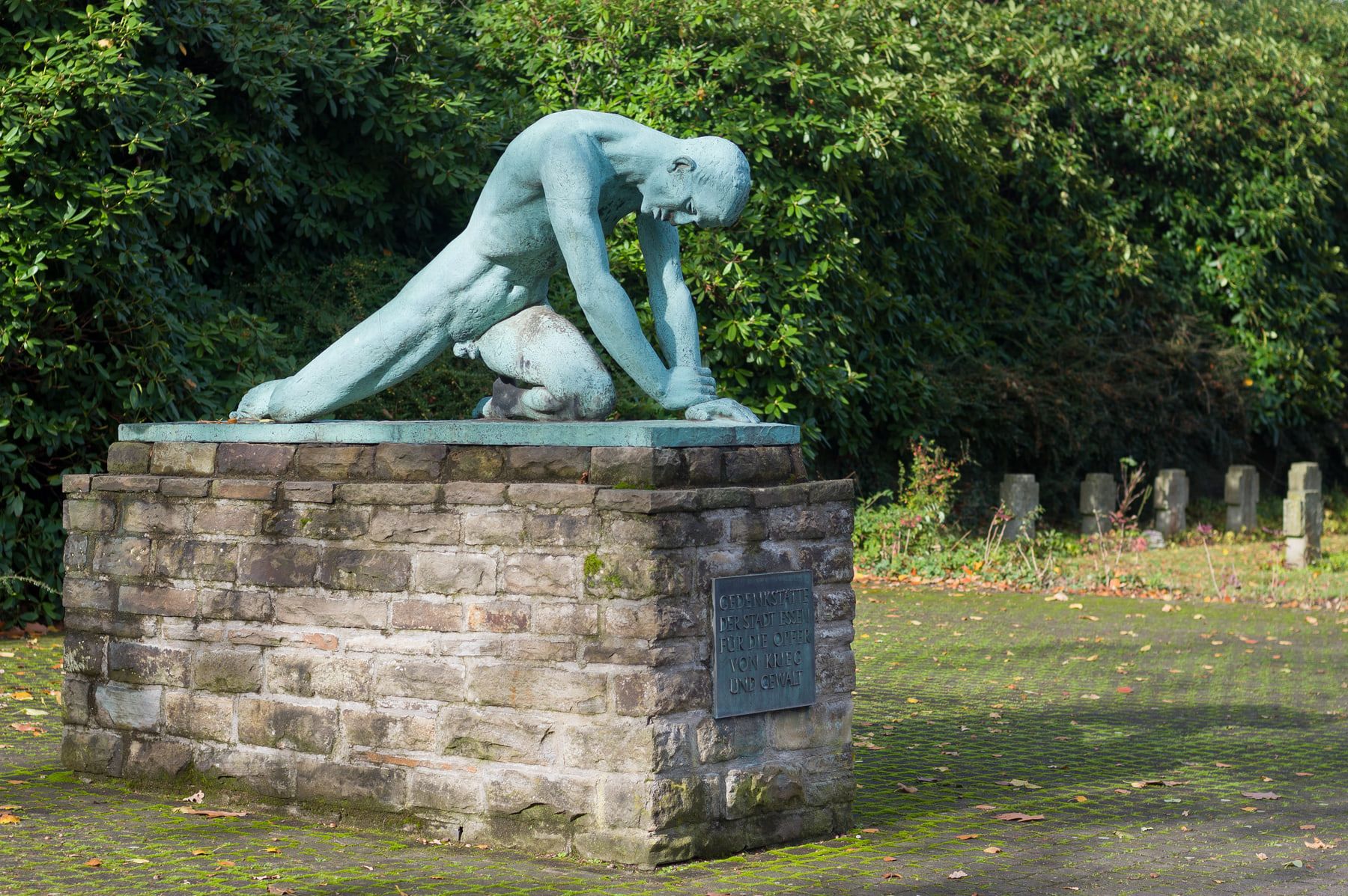 Joseph Enselings grün patinierte Skulptur „Trauer“ mit einer tief gebeugten Figur auf einem gemauerten Sockel; dahinter sind Gehölze und Grabsteine zu sehen. auf Südwestfriedhof Essen