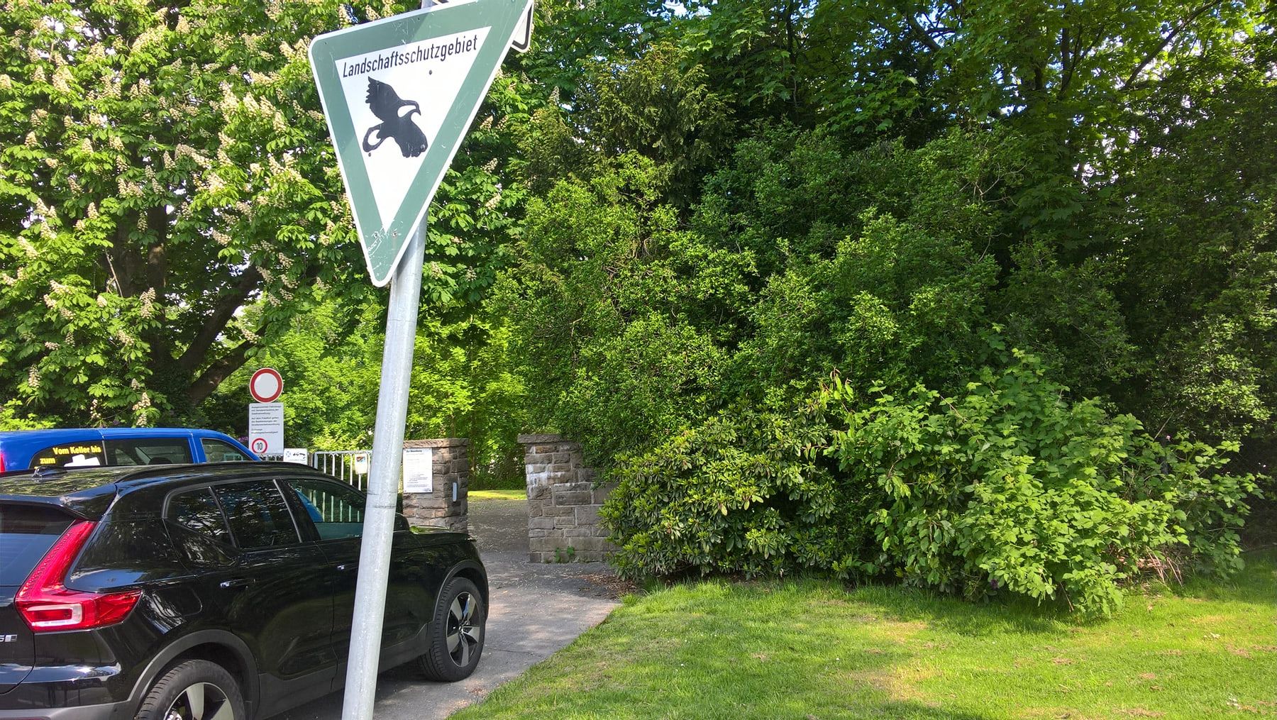 Von parkenden Autos und einem Schild „Landschaftsschutzgebiet“ teilweise verdeckter Friedhofseingang zwischen niedrigen Natursteinpfeilern auf Südwestfriedhof Essen